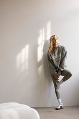 Model wearing Trui Academie Vetiver sweater in vetiver green, styled with leggings and sneakers against a minimalist background.