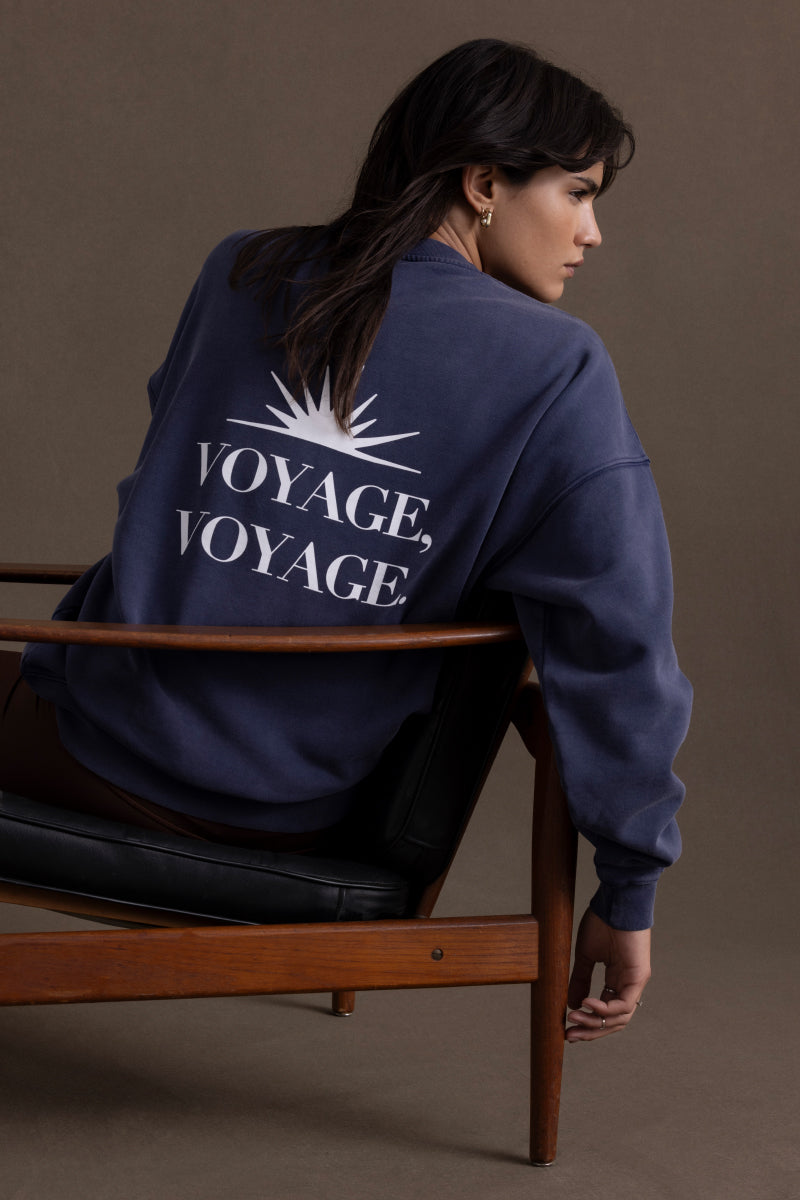 Woman in navy sweater with "Voyage, Voyage" print sitting on chair, showcasing modern style and relaxed fit.