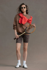 Model wearing a cocoa sweater with a red accessory, paired with leopard print shorts, holding a tennis racket.
